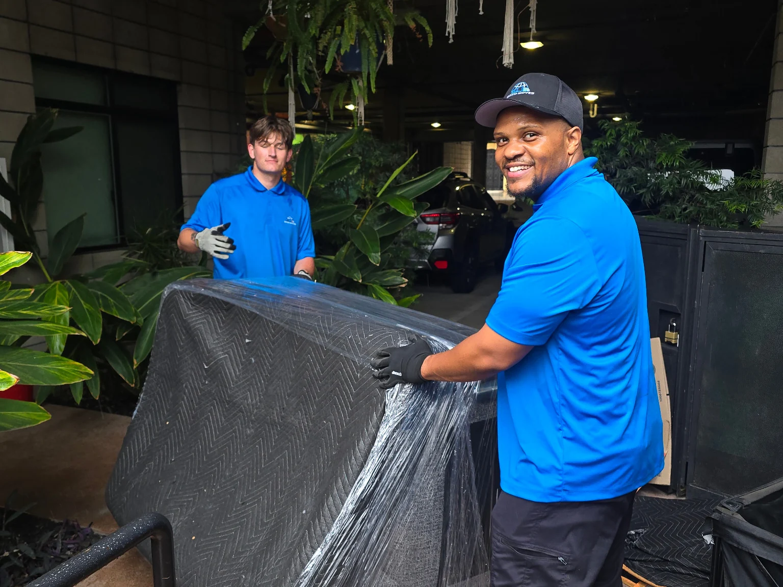 Two uniformed Soco Moves crew members handling blanket-wrapped furniture in a covered courtyard.